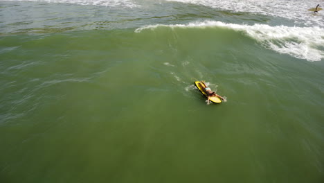 una toma aérea de una surfista tendida en una tabla de surf amarilla y nadando en dirección a una ola