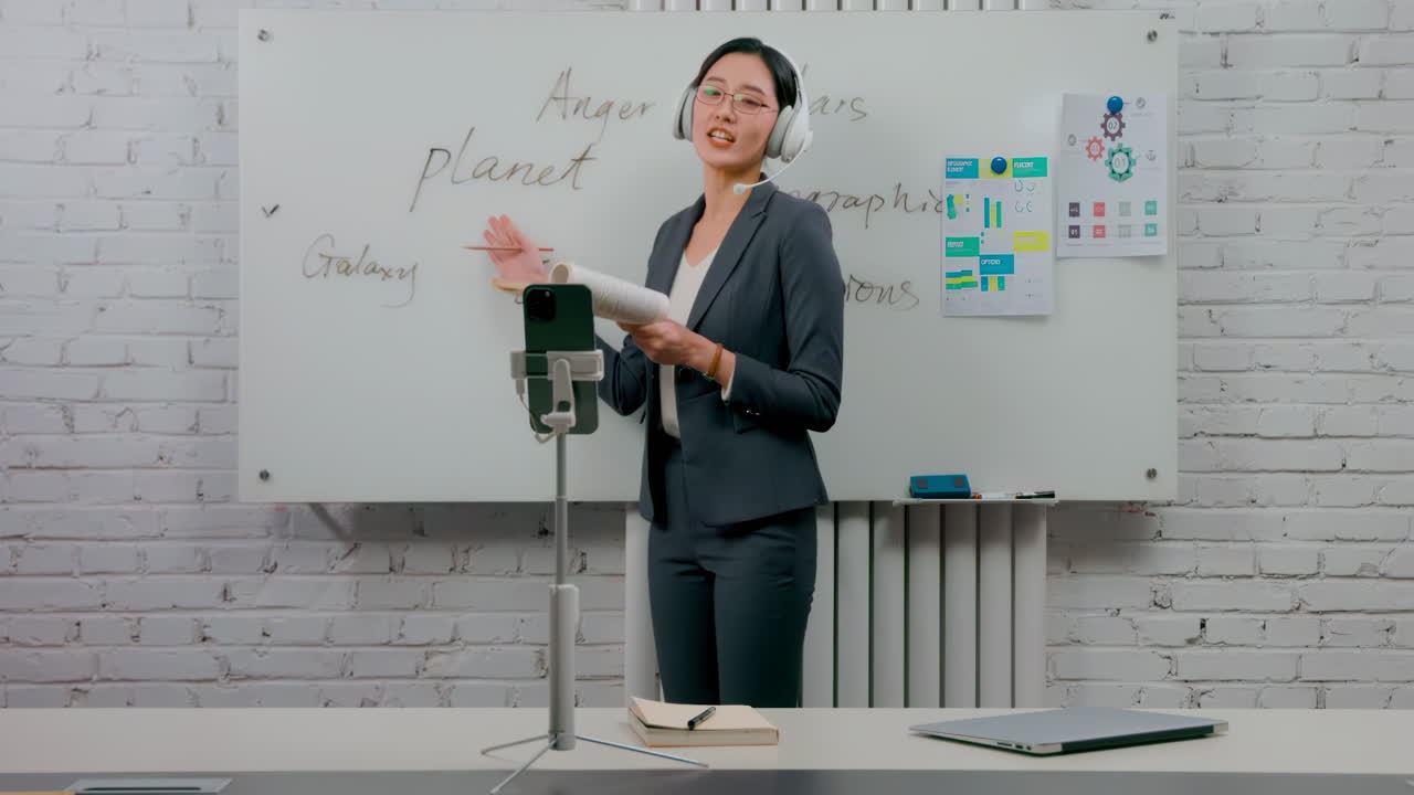 Portrait of self-confident businesswoman glasses standing in the office at the desk with a headset, coach with paperwork and graphs, explaining information in front of cell phone camera