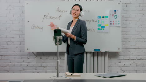 portrait of self-confident businesswoman glasses standing in the office at the desk with a headset, coach with paperwork and graphs, explaining information in front of cell phone camera