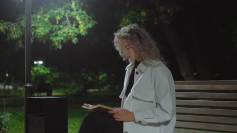 woman seated in solitude at night reading book under streetlight on park bench as wind gently moves nearby tree leaves and creates soft glow in quiet atmosphere