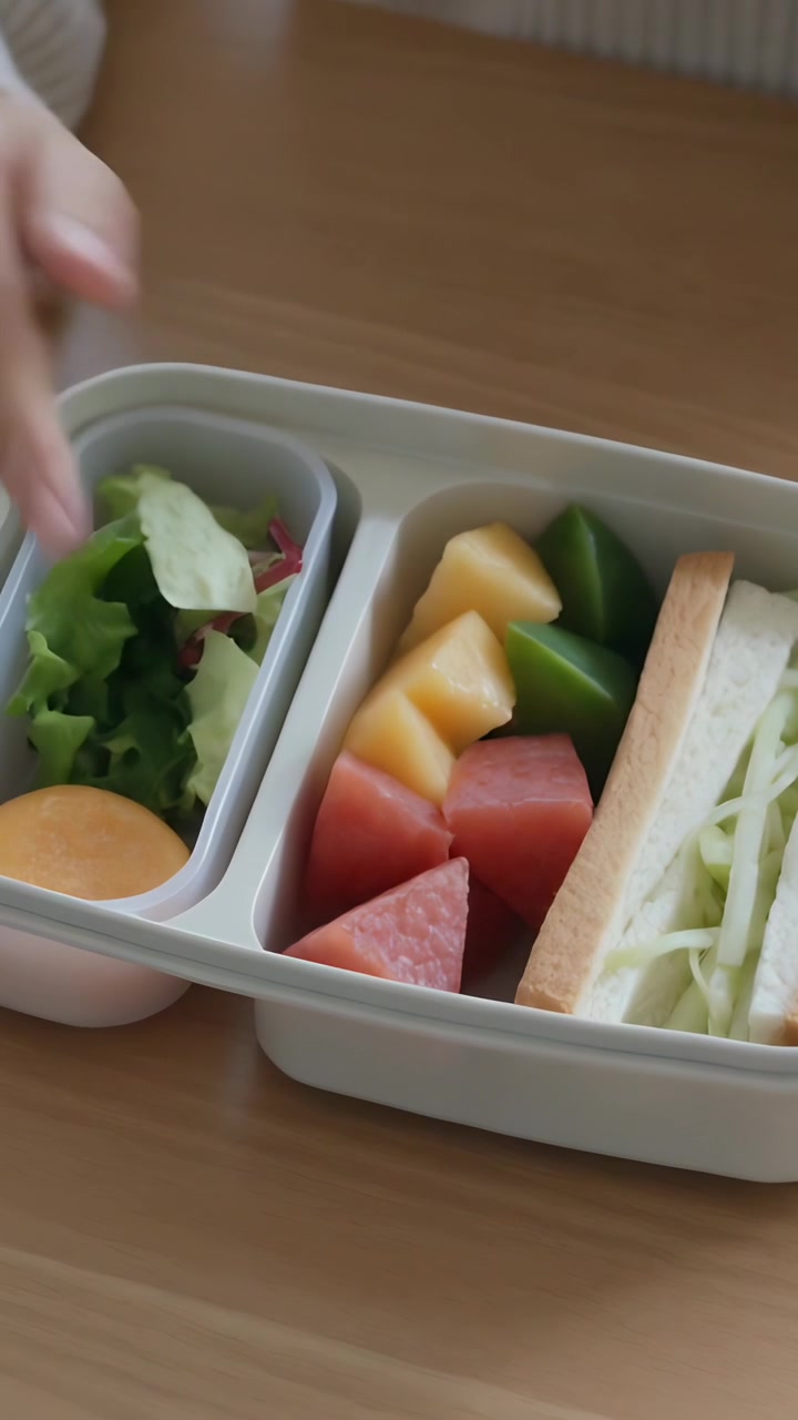 Vertikales Video: Frau legt Orange in Bento-Salat auf Tisch, gerippter Ärmel.