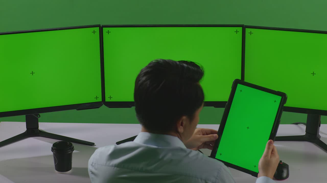 태블릿과 여러 컴퓨터 모니터로 작업하는 아시아 남자의 뒷면은 녹색 스크린 스튜디오에서 녹색 화면 모형을 가지고 있습니다.