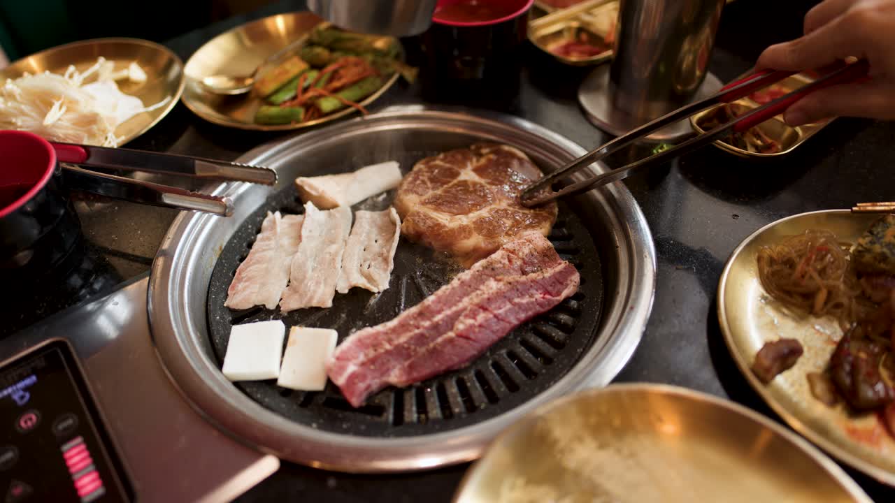 Hand places pork belly on tabletop grill with side dishes in warm, inviting restaurant lighting