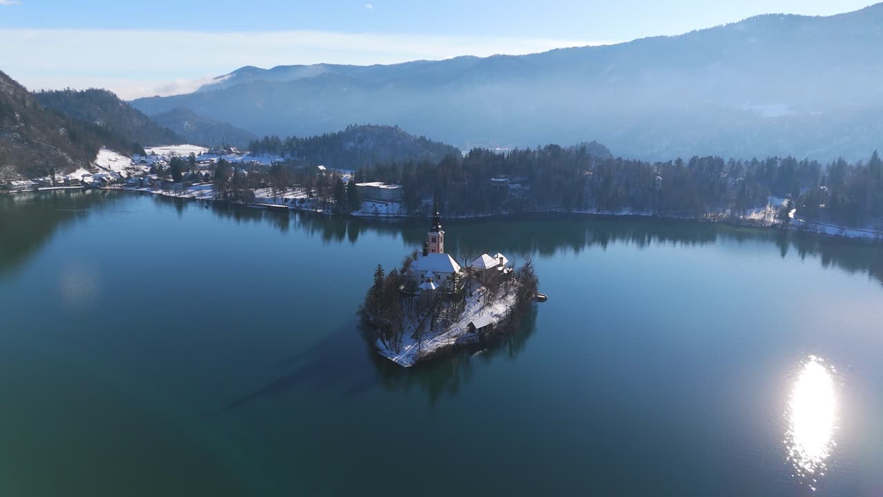 Video aereo con dolly che mostra l'iconica Isola di Bled e la chiesa sul Lago di Bled, Slovenia, durante una fredda giornata invernale. Paesaggio montano scenografico con alberi coperti di neve e acqua blu calma.