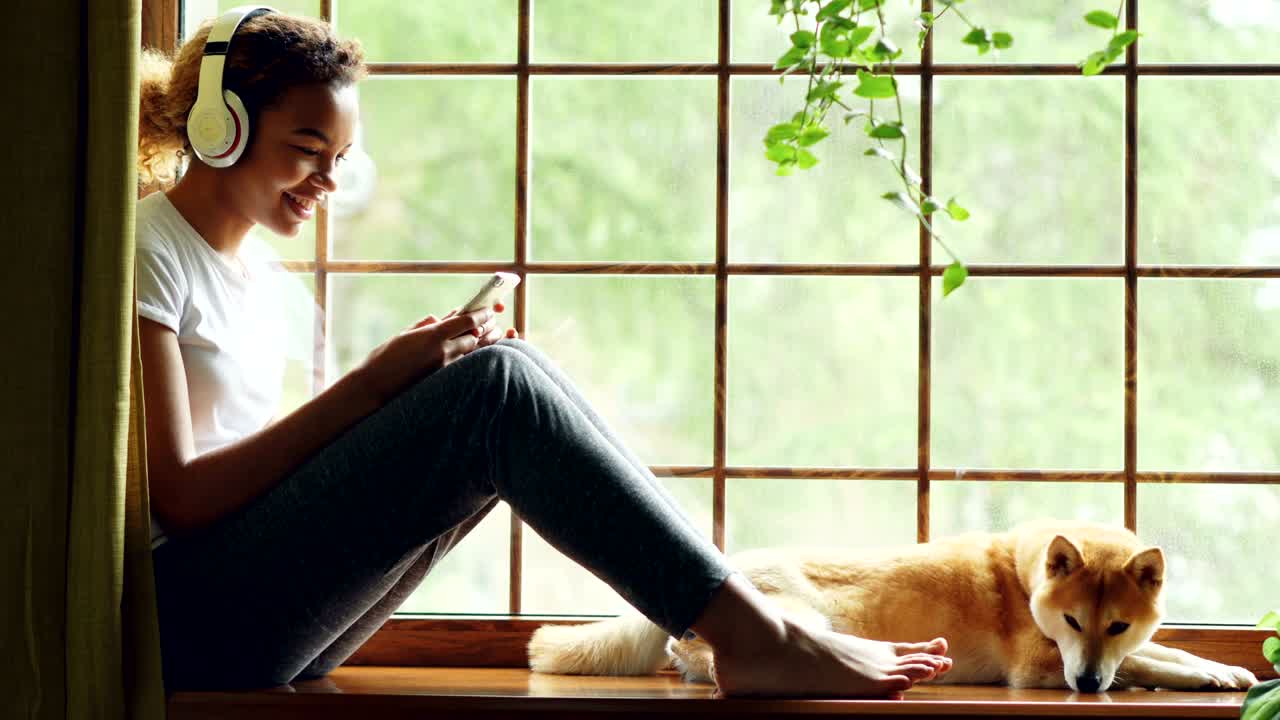 Attractive young woman is listening to music through wireless headphones and choosing song on smartphone, her pet dog is lying on windowsill near its owner.