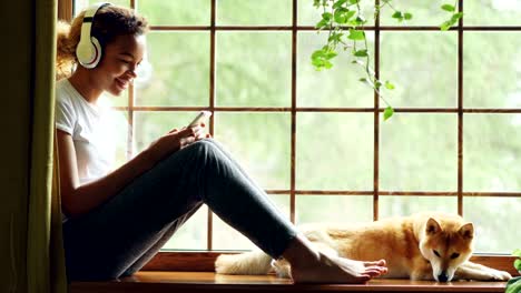 attractive young woman is listening to music through wireless headphones and choosing song on smartphone, her pet dog is lying on windowsill near its owner.