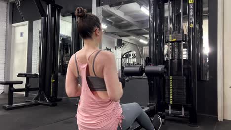 a woman pulling weights towards herself with equipment in the gym is exercising, woman with glasses is doing sports