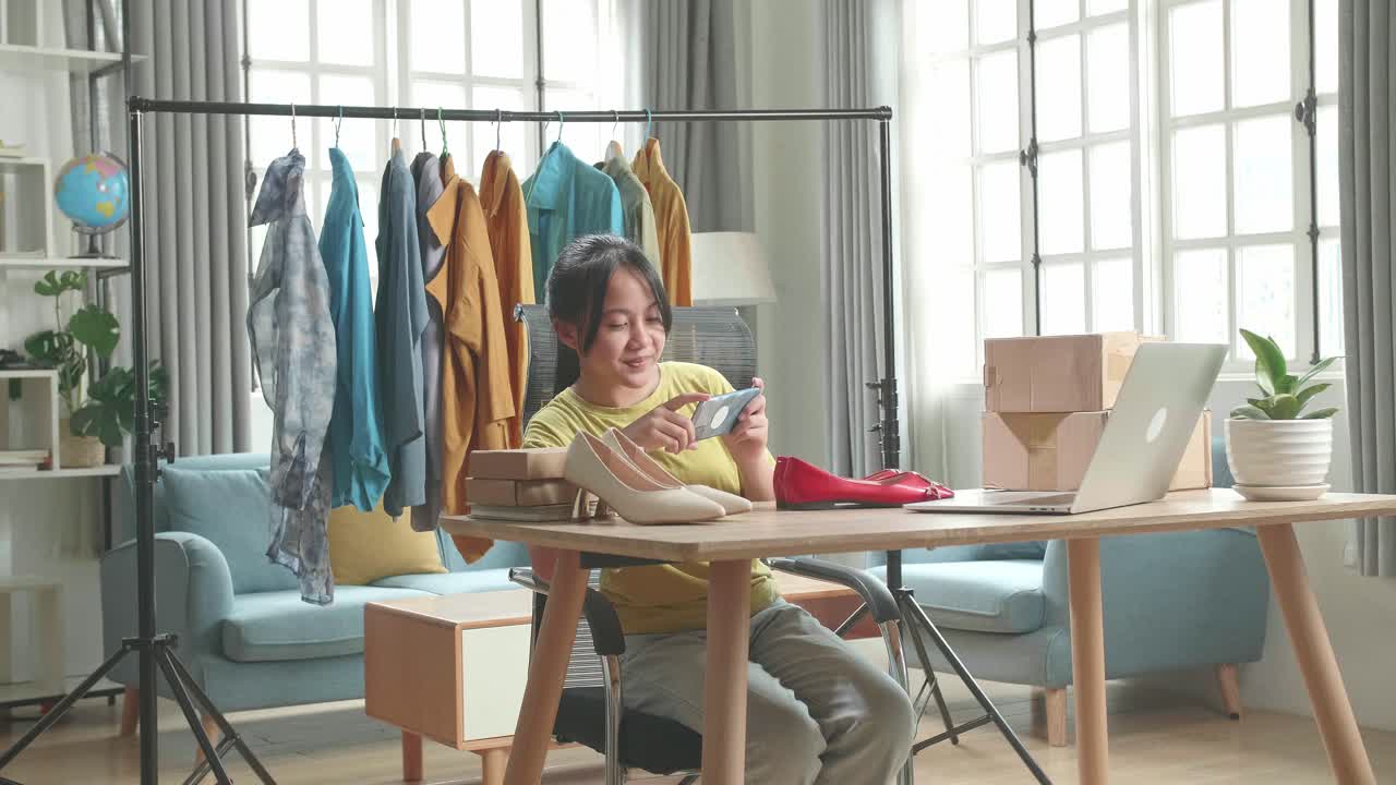 Asian Young Girl Online Seller Taking Photos With Mobile Phone While Using Computer For Selling Clothes At Home