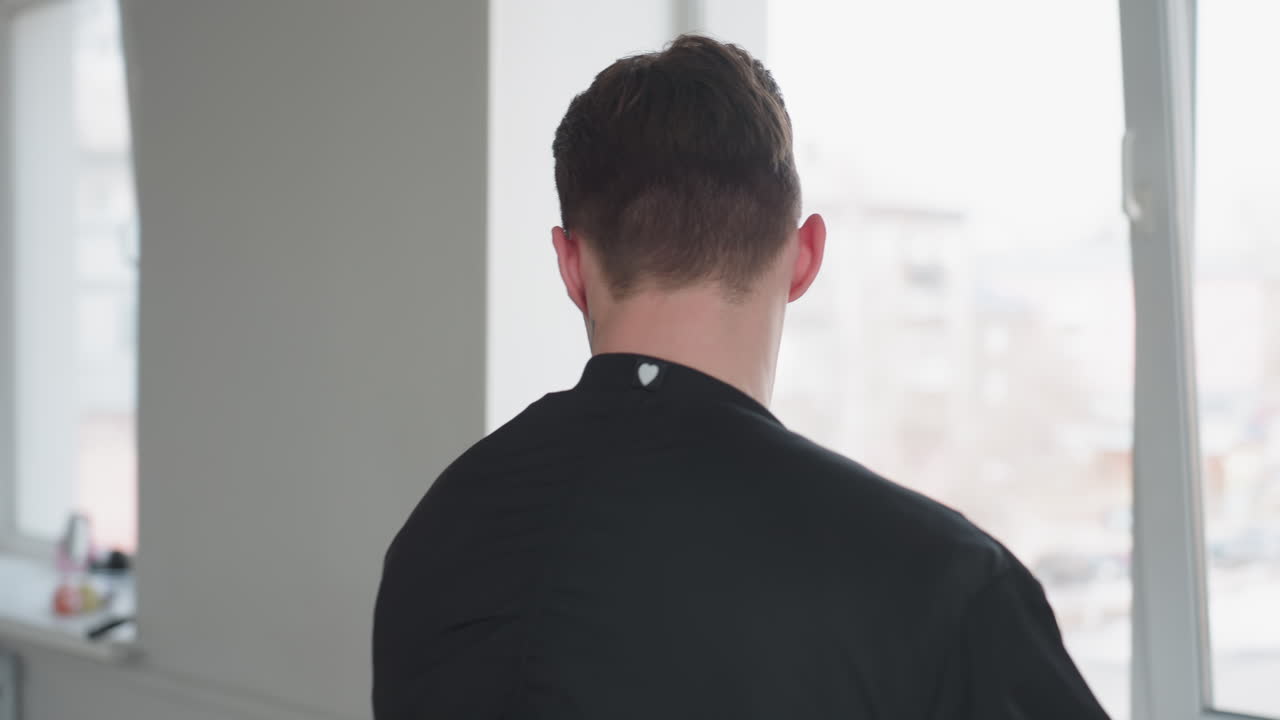 Young man in black jacket walks toward window while pushing suitcase handle inward, blur of outdoor scene through window in background