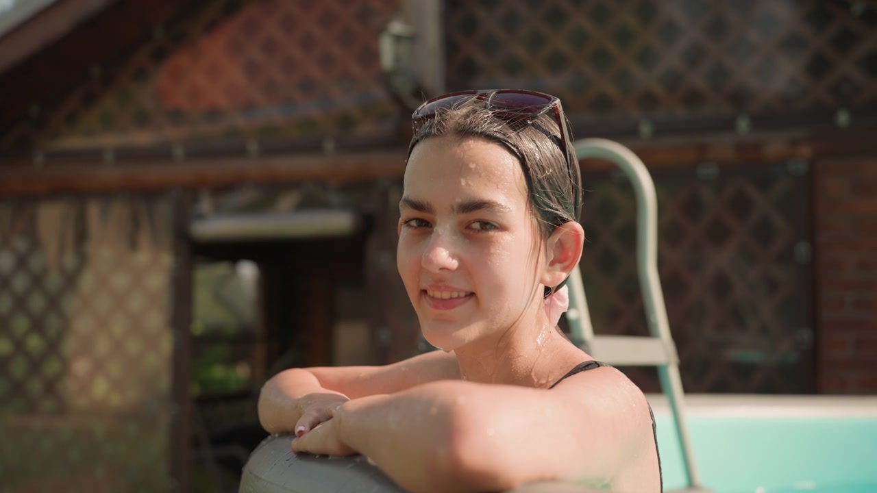 Young woman relaxes in small inflatable pool outdoors adjusting her hair and enjoying water. Woman is wet with glistening skin against outdoor backdrop featuring greenery and wooden