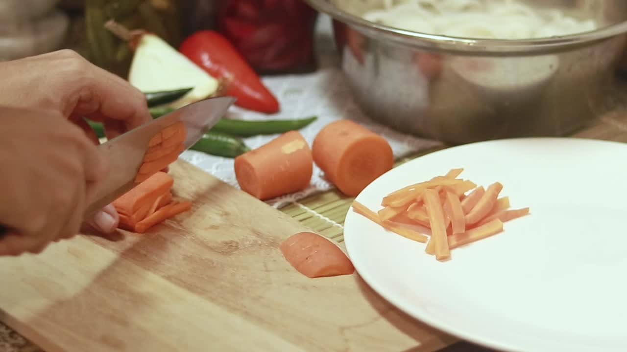 Slicing raw carrots into thin strips on a wooden chopping board and setting it aside on a plate, preparing ingredients for cooking
