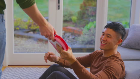 man surprising same sex partner at home with heart shaped gift held behind his back