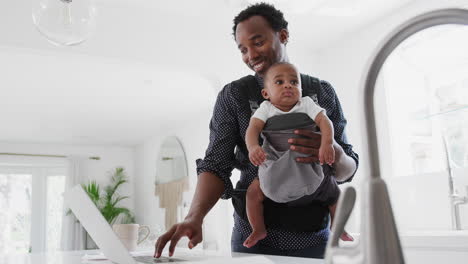 Padre-Con-Hijo-En-Cabestrillo-Multitarea-Trabajando-Desde-Casa-En-Una-Computadora-Portátil