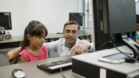 Teacher-explaining-schoolgirl-how-computer-works-and-typing-on-keyboard-while-girl-listening-attentively