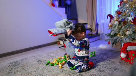 dark-haired baby boy in winter pajama sits near christmas tree. lovely boy gets some candies from the stocking.