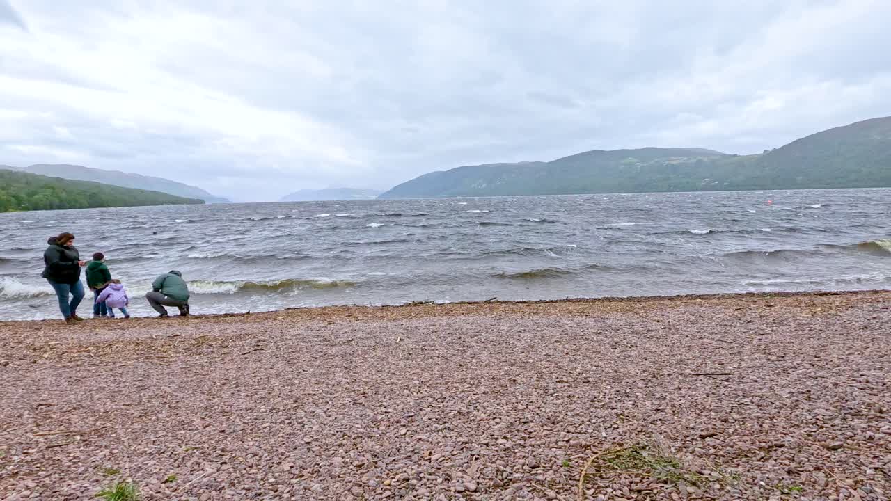 Group of people walking from grassy path to pebbled lakeshore under overcast summer sky