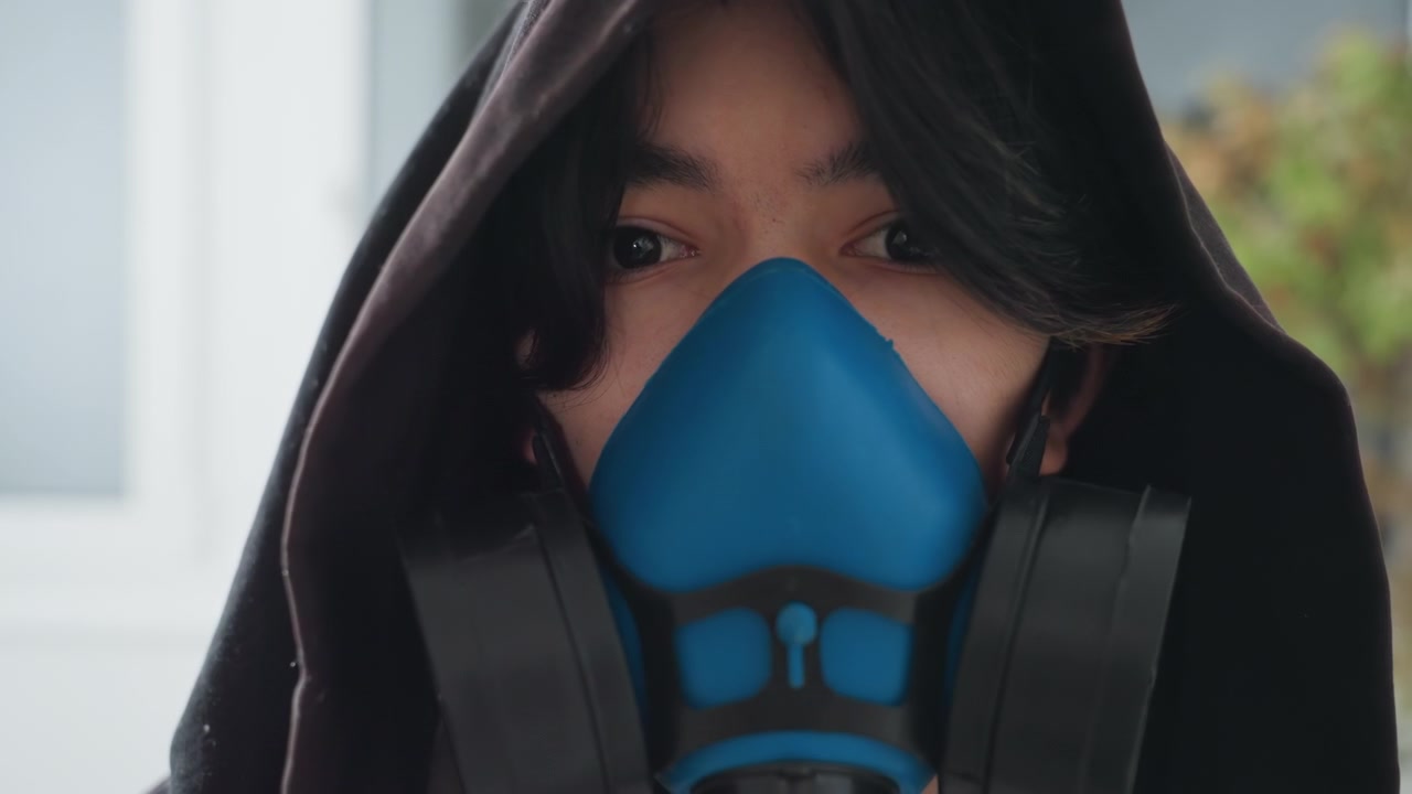 Gemaskerde schoonmaker in huis draagt ademhalingsmasker, technicus met capuchon inspecteert binnenraam, blauw ademhalingsmasker en filters zichtbaar, handschoenen geïmpliceerd, gerichte blik, scène van huishoudelijke sanering.