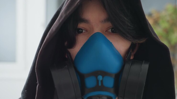 Masked cleaner in home wearing respirator, hooded technician inspects indoor window area, blue respirator facepiece and filters visible, gloves implied, focused gaze, domestic remediation scene