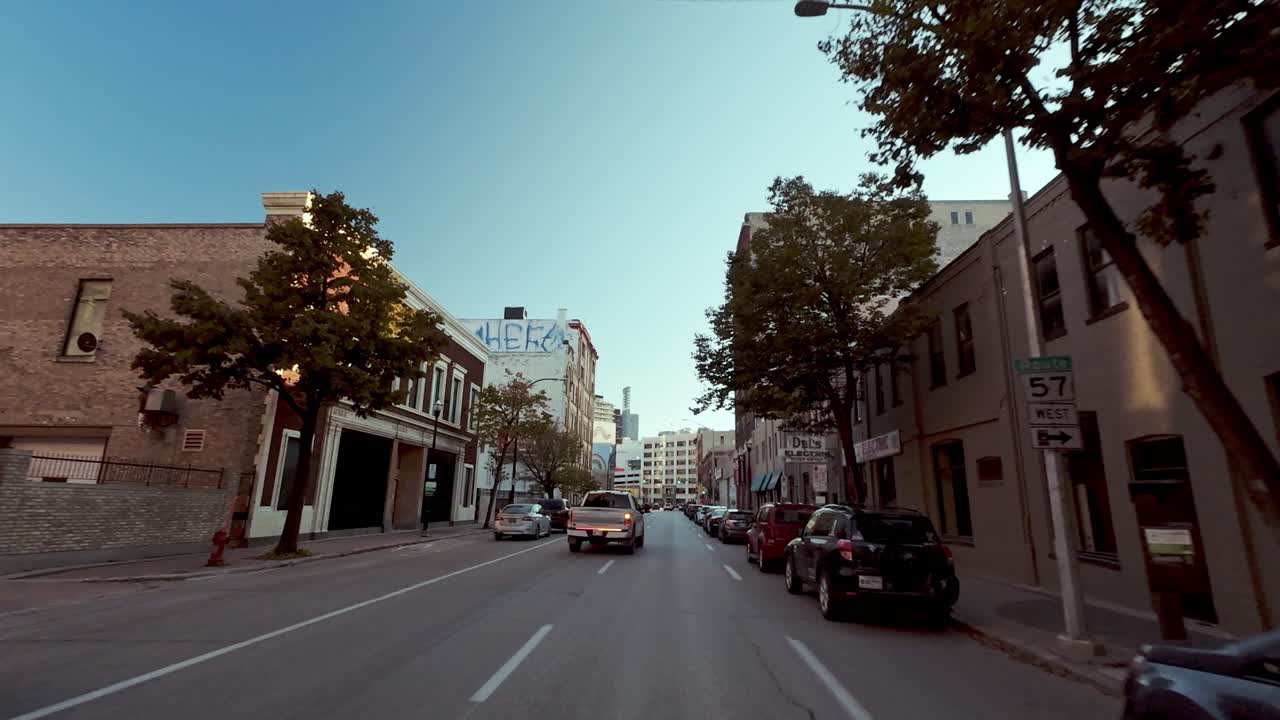 POV shot from car driving down street in older American city; concrete and brick buildings, vintage ads, blue sky