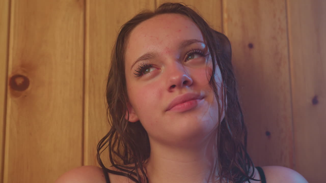 two women wrapped in towels relax against wooden sauna wall with soft smoky steam swirling, damp hair resting on shoulders, ambient light casting warm glow, eyes closed in tranquil moment