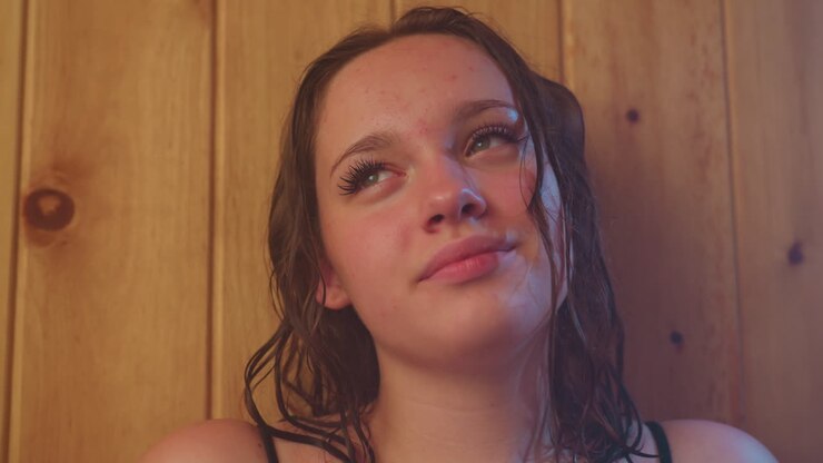 two women wrapped in towels relax against wooden sauna wall with soft smoky steam swirling, damp hair resting on shoulders, ambient light casting warm glow, eyes closed in tranquil moment