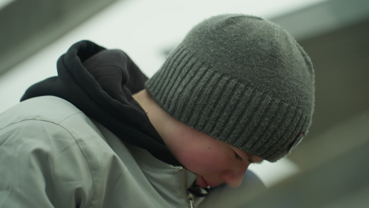 Close-up of a young boy wearing a gray jacket and green beanie, deeply focused on a task near an iron railing, he looks frustrated