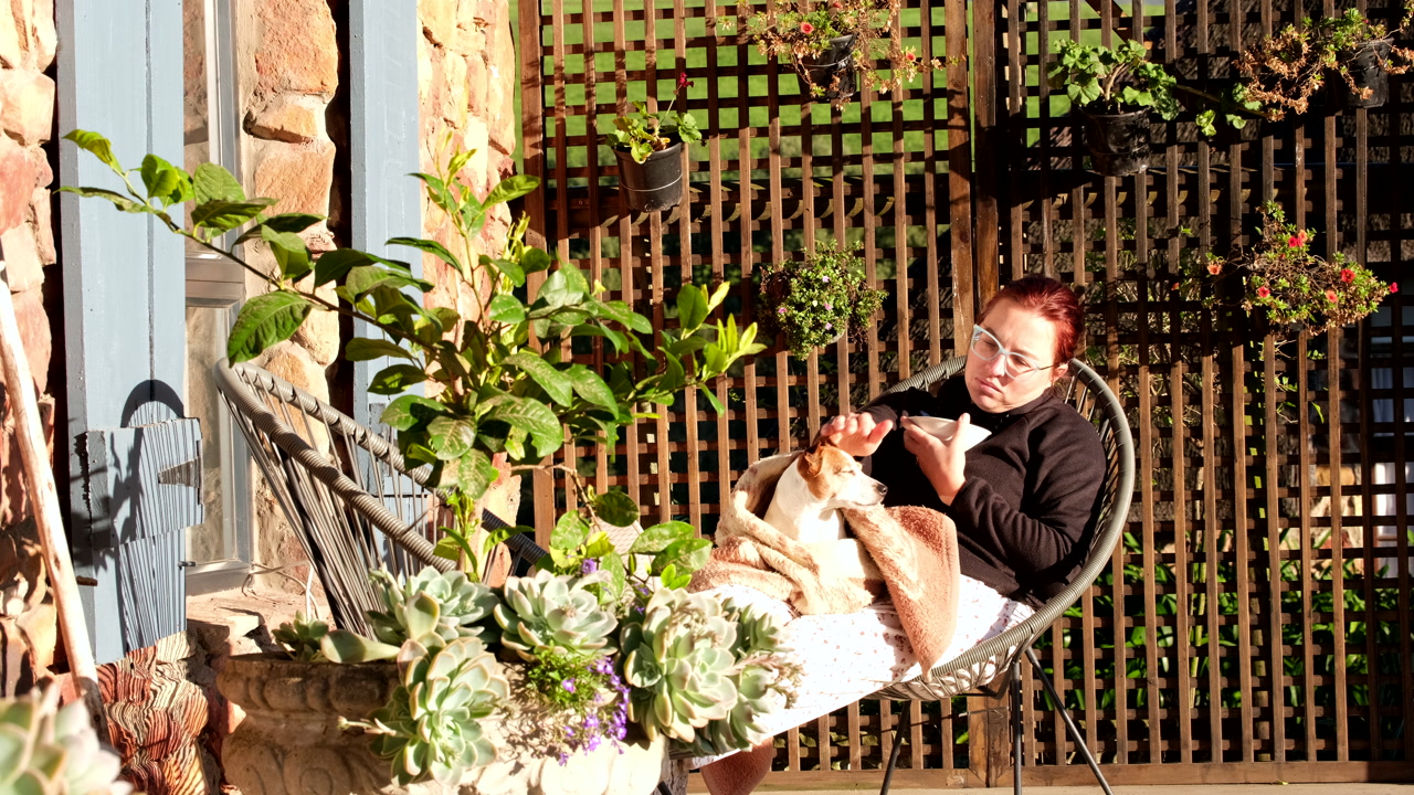 Woman with pet dog on lap sits on farm house terrace eating cereal at sunrise