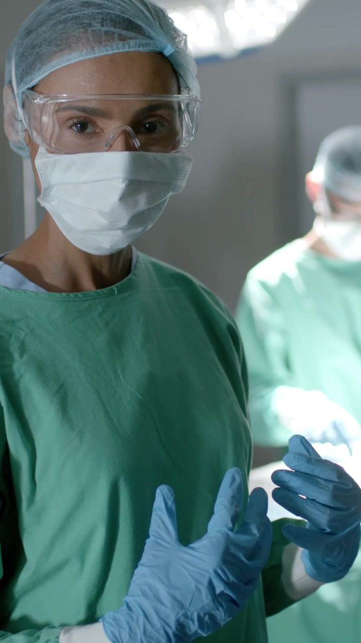 Vídeo vertical: Equipe cirúrgica, cirurgiã ajustando luvas azuis antes da cirurgia, garantindo a esterilidade.