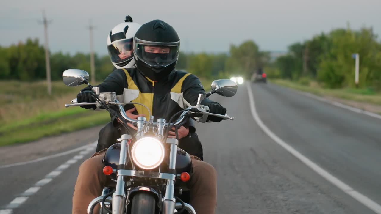 Man and woman wearing helmets ride motorcycle on morning countryside trip, driving along asphalt road with car passing on opposite lane carrying load, surrounded by trees, and grassy roadside