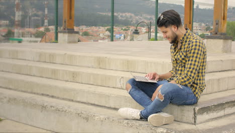 Hübscher-Kerl,-Der-Auf-Treppen-Sitzt-Und-An-Seinem-Laptop-Und-Handy-Arbeitet