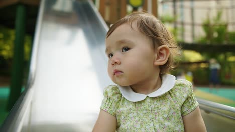 cute little one-year-old baby girl sitting on the slider and staring at something with curiosity and astonishment, spying facial expression