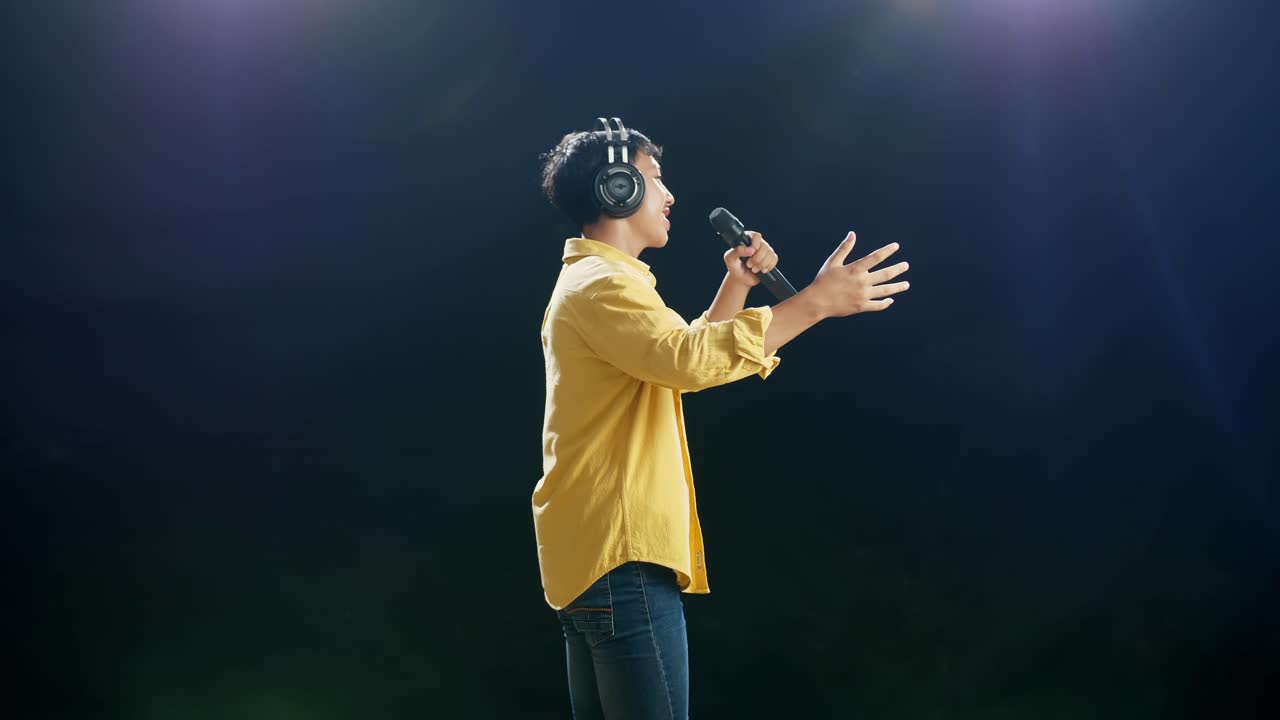 vista lateral de un joven asiático con auriculares sosteniendo un micrófono, bailando y cantando en el fondo negro