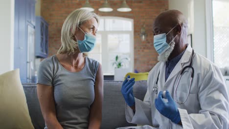 happy senior diverse woman and doctor wearing face masks in living room sitting on sofa, vaccinating