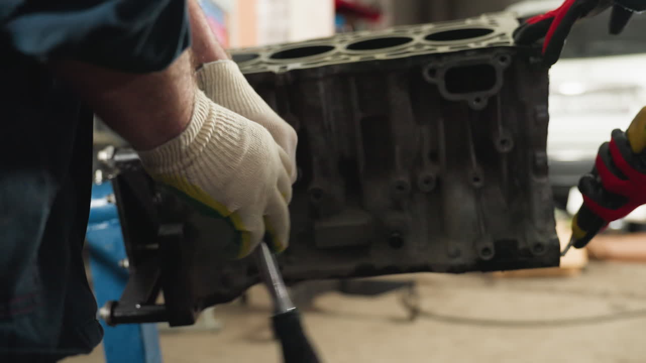 primer plano de un mecánico en uniforme azul usando un raspador amarillo en el motor del coche mientras otro mecánico con guantes rojos ayuda, trabajando juntos en un taller automotriz ocupado con herramientas y equipos