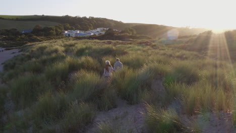 Drone-Shot-Of-Senior-Couple-Holding-Hands-As-They-Walk-Across-Dunes-On-Winter-Beach-Vacation