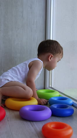 little kid sits on the floor with toy pyramid scattered around. toddler looks at window leaning on the glass with his head. side view. vertical video