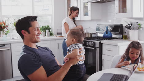 Familia-Hispana-Hablando-En-Su-Cocina,-Mamá-Cocinando-En-La-Hornilla,-Papá-Levantando-Al-Bebé-En-El-Aire,-De-Cerca