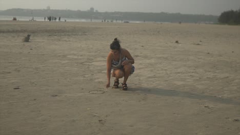 mujer india en un vestido floral se agacha para dibujar en la arena en una playa tranquila