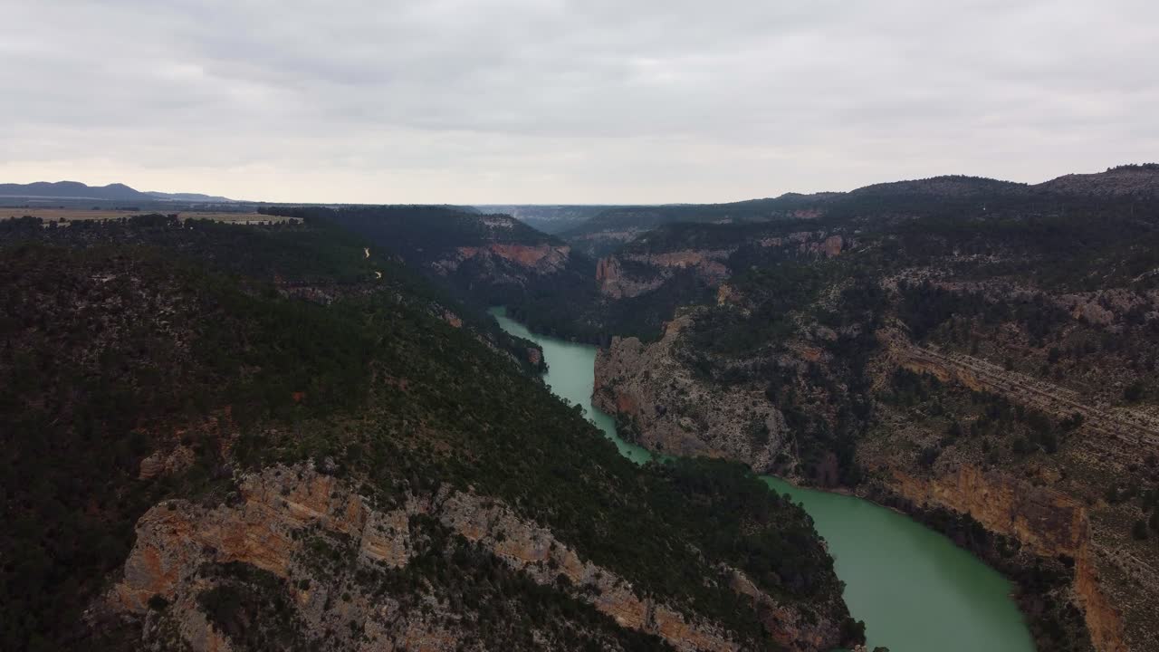 Bredt 4K luftpanorama over den svingete Júcar-elvens canyon i Spania, med tette furuskoger, robuste kalksteinsklipper og en vidstrakt fjellhorisont under en skyet himmel