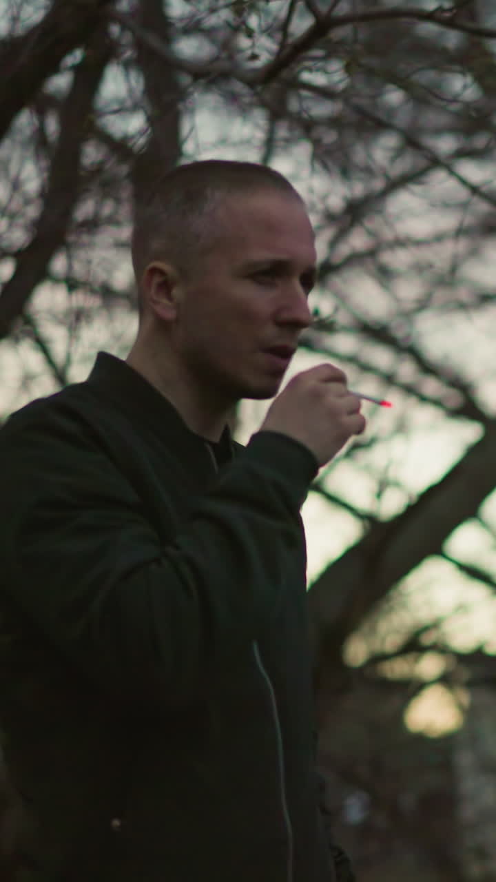 una vista lateral de un hombre con una chaqueta verde fumando un cigarrillo en el bosque al anochecer, con el fondo borroso de los árboles y la luz oscurecida