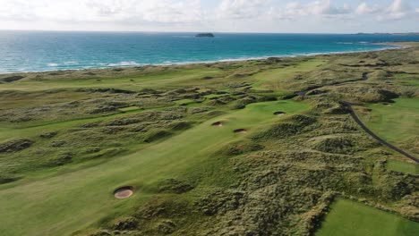 drone flies over green rolling fairway of ireland links golf course along coastline with blue waves crashing