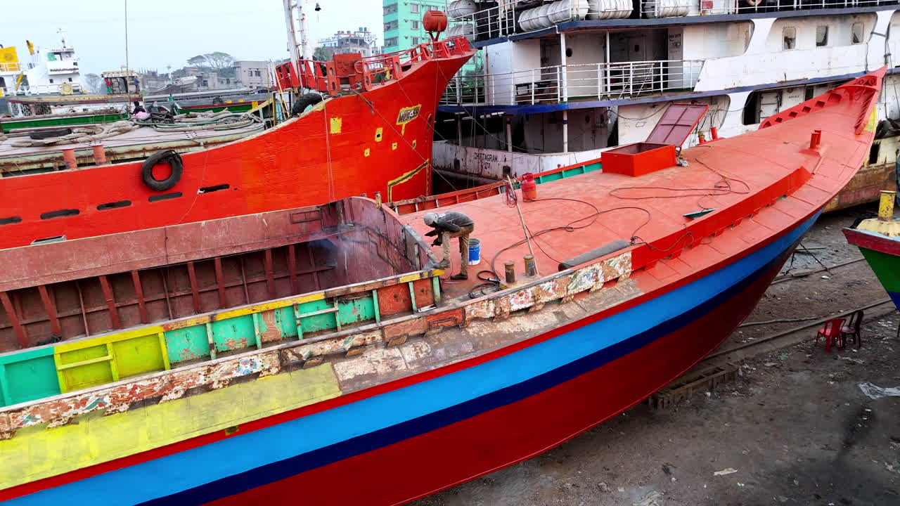 Cinematic slow circling aerial shot of a welder working on a cargo ship docked on the shore of the Buriganga river in Dhaka, Bangladesh.