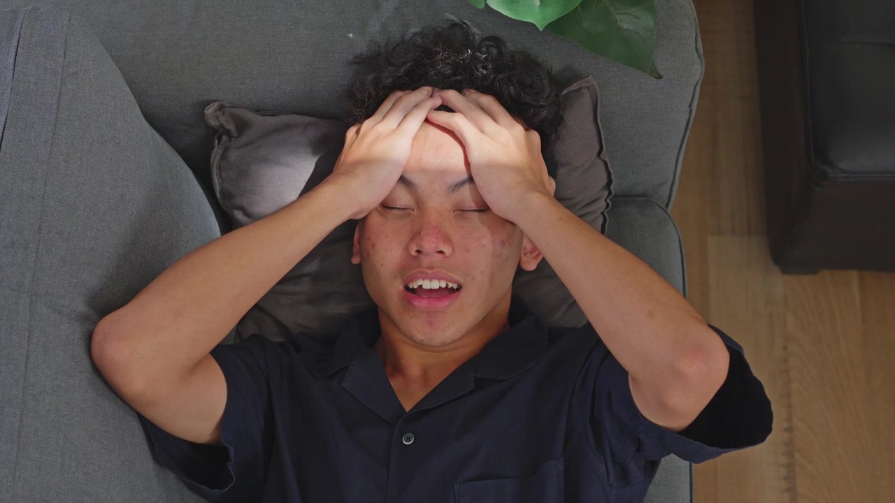 Top view of young Asian man lying on couch grimacing in pain clutching head from severe migraine