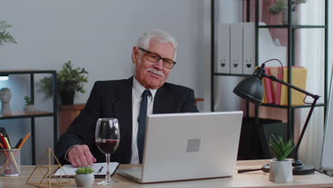 Senior-man-freelancer-drinking-wine-after-working-on-laptop-computer-pc-sitting-at-desk-at-office