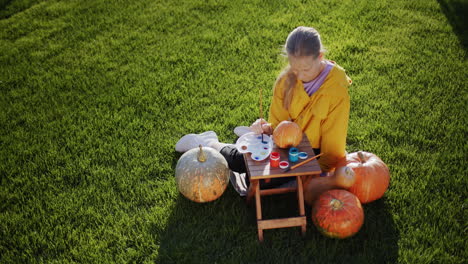 Top-view:-A-child-paints-a-pumpkin,-prepares-decorations-for-Halloween.-Sits-on-the-lawn-in-the-backyard-of-the-house