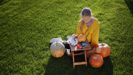 top view: a child paints a pumpkin, prepares decorations for halloween. sits on the lawn in the backyard of the house