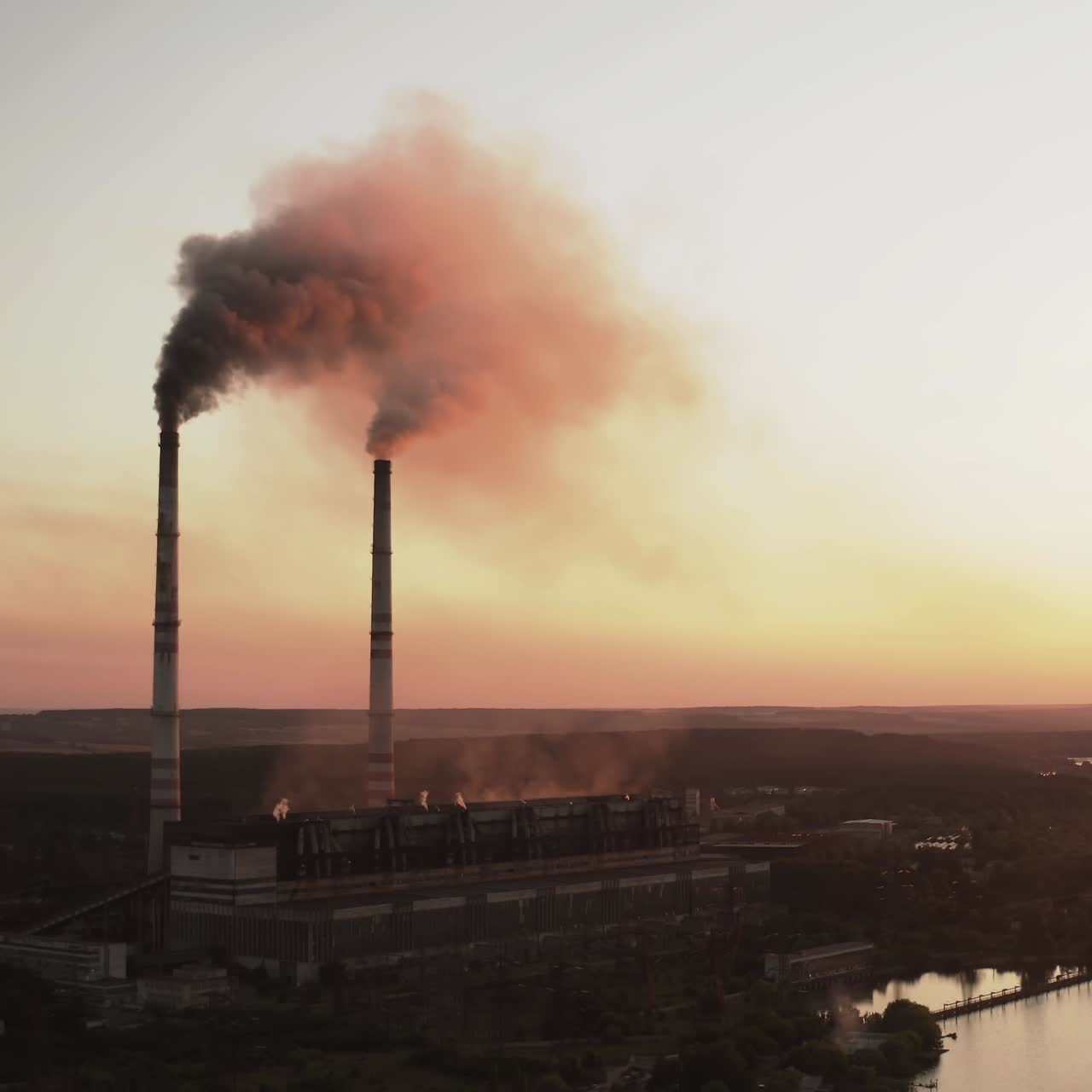 Harmful factory near the river at dusk. Industrial zone with chimneys pouring dark smoke into the air at sunset. Pollution of the environment. Aerial view