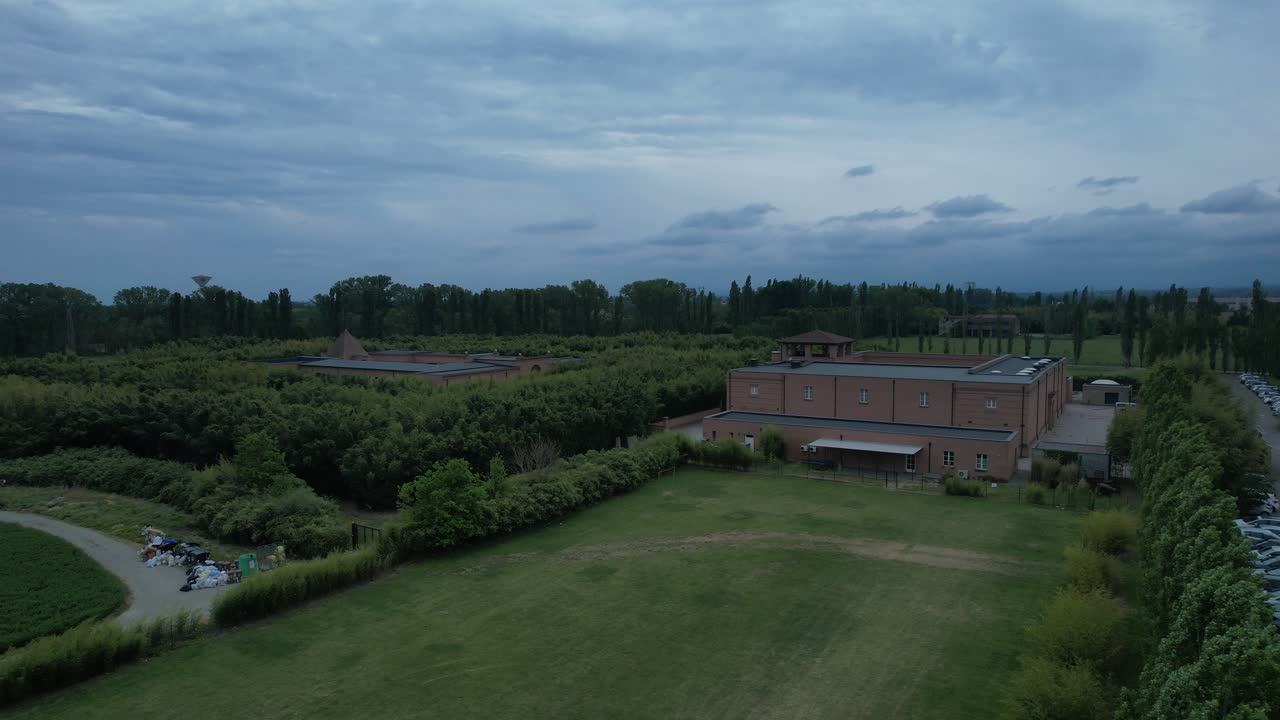 Il Labirinto della Masone, un labirinto di bambù a Fontanellato, provincia di Parma, Italia, che mostra il labirinto geometrico, gli edifici museali, le siepi verdi e il paesaggio pianeggiante della Pianura Padana sotto un cielo nuvoloso, ripresa con drone in discesa.