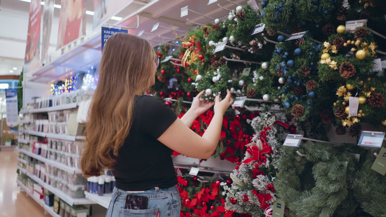 vista lateral de una mujer comprando decoraciones navideñas en una tienda minorista, examinando coronas festivas adornadas con adornos, piñas y cintas, estantes llenos de coloridas decoraciones estacionales