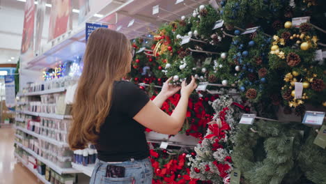 side view of woman shopping for holiday decorations in retail store, examining festive wreaths adorned with ornaments, pinecones, and ribbons, shelves filled with colorful seasonal decor